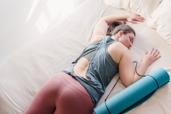 A woman in activewear resting on a bed with a yoga mat, symbolizing relaxation and fitness.