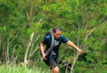 A woman carries a tire outdoors during a fitness challenge surrounded by greenery.