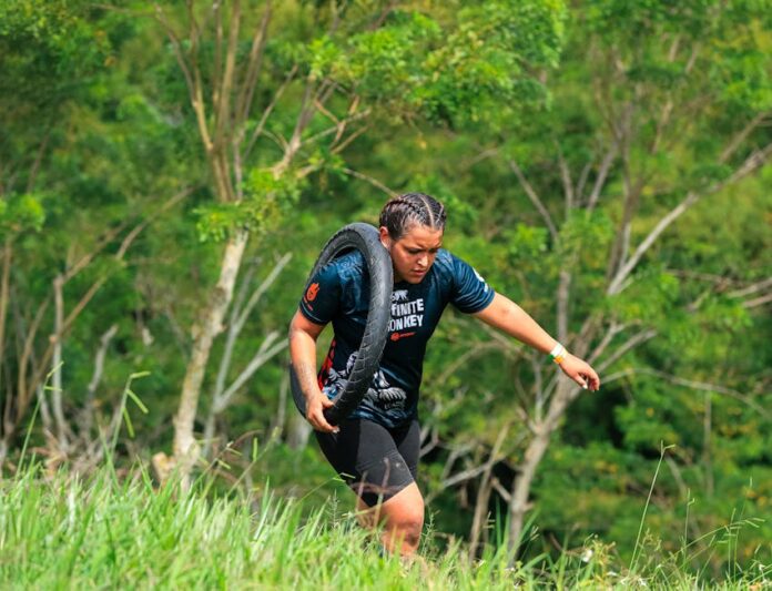 A woman carries a tire outdoors during a fitness challenge surrounded by greenery.