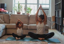 Full body content fit Asian woman and daughter in activewear sitting in Lotus Pose with arms raised and looking at each other while practicing yoga in cozy living room