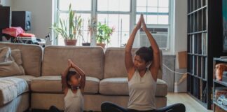 Full body content fit Asian woman and daughter in activewear sitting in Lotus Pose with arms raised and looking at each other while practicing yoga in cozy living room