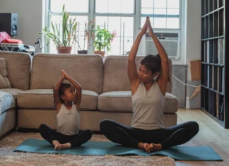 Como criar uma rotina diária de exercícios em casa facilmente Full body content fit Asian woman and daughter in activewear sitting in Lotus Pose with arms raised and looking at each other while practicing yoga in cozy living room