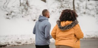 Back view unrecognizable ethnic sporty male trainer and plump female in outerwear jogging together on snowy roadside on cold winter weather