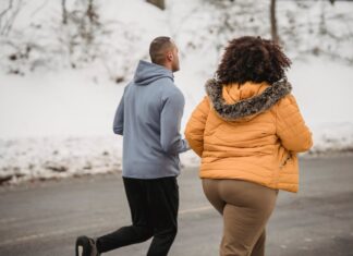 Back view unrecognizable ethnic sporty male trainer and plump female in outerwear jogging together on snowy roadside on cold winter weather