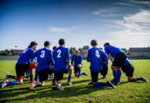 A group of male football players in blue uniforms huddle on a grassy field before a match.