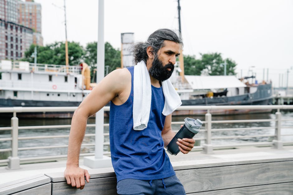 A man with a towel and water bottle rests by the waterfront after exercise.