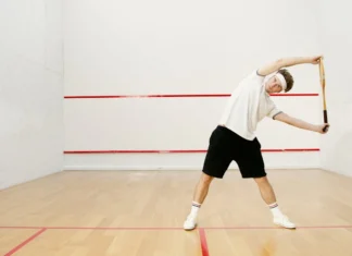 Como treinar a resistência para jogar Squash de forma eficaz Young man stretching with a racket on an indoor squash court, preparing for a workout.