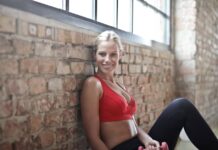 A fit woman in a red sports bra smiling while sitting against a brick wall after a workout.