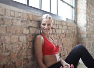 A fit woman in a red sports bra smiling while sitting against a brick wall after a workout.