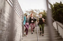 Emagrecer de forma saudável: Caminhada diária pode transformar vidas Three women walking down steps outdoors, carrying yoga mats, enjoying a fitness conversation.
