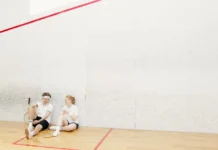 Two young squash players resting on a court, holding rackets and chatting.