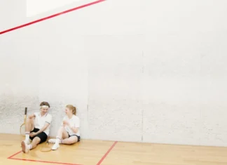 As melhores raquetes de Squash para amadores Two young squash players resting on a court, holding rackets and chatting.