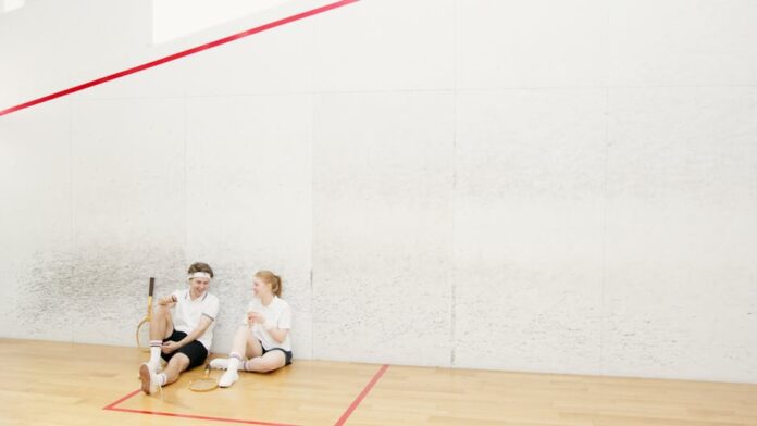 Two young squash players resting on a court, holding rackets and chatting.