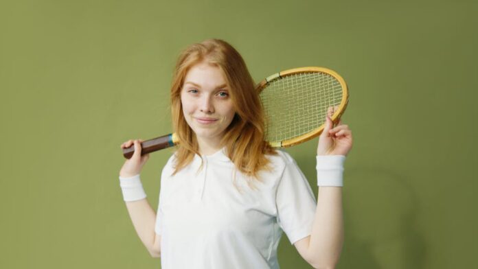Portrait of a young caucasian woman holding a squash racket against a green background.