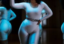Woman in white leotard posing confidently in front of gym mirror, showcasing fitness and style.