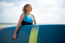 A senior woman in a wetsuit holding a surfboard, enjoying a sunny day on the Portuguese coast.