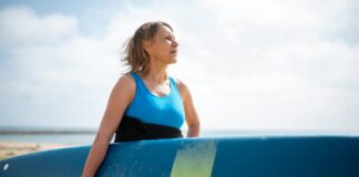A senior woman in a wetsuit holding a surfboard, enjoying a sunny day on the Portuguese coast.