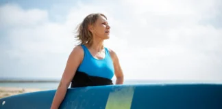 A senior woman in a wetsuit holding a surfboard, enjoying a sunny day on the Portuguese coast.