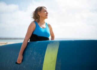 A senior woman in a wetsuit holding a surfboard, enjoying a sunny day on the Portuguese coast.