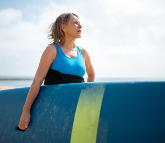 Como aprender Surf em Portugal: Dicas imperdíveis para iniciantes A senior woman in a wetsuit holding a surfboard, enjoying a sunny day on the Portuguese coast.