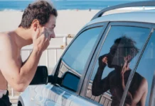 A man applies sunscreen using his car window reflection at a sunny beach.