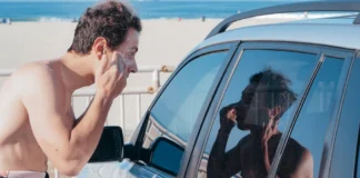 A man applies sunscreen using his car window reflection at a sunny beach.