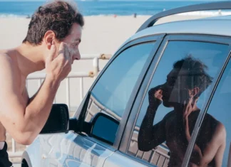 Cuidados de segurança para surfistas iniciantes: Dicas essenciais A man applies sunscreen using his car window reflection at a sunny beach.