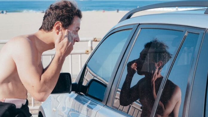 Cuidados de segurança para surfistas iniciantes: dicas essenciais! A man applies sunscreen using his car window reflection at a sunny beach.