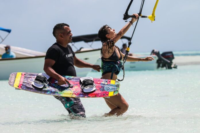 Melhores praias para iniciantes no surf: Descubra as ideais! Man assisting woman with kitesurfing on a sunny tropical beach, enjoying the thrill and recreation.