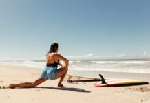 Woman stretching on beach with surfboard ready, under clear blue sky.