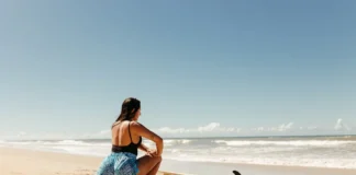 Treinos rápidos para fazer entre as refeições de Natal e emagrecer Woman stretching on beach with surfboard ready, under clear blue sky.
