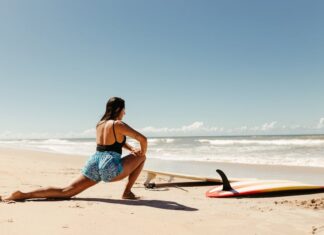 Woman stretching on beach with surfboard ready, under clear blue sky.