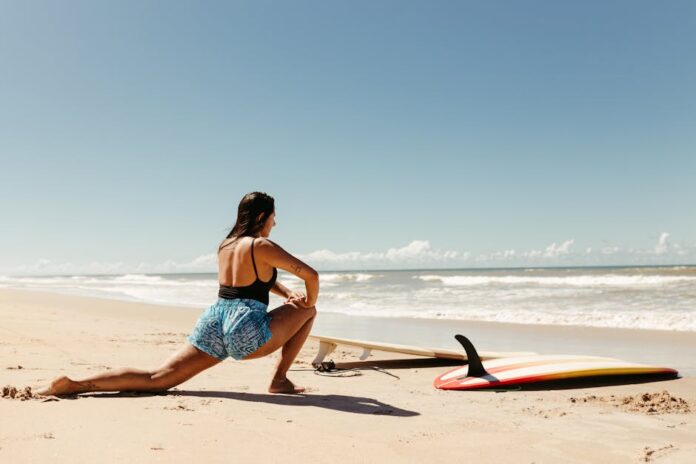 Treinos Rápidos para Fazer Entre Refeições de Natal e Emagrecer Woman stretching on beach with surfboard ready, under clear blue sky.