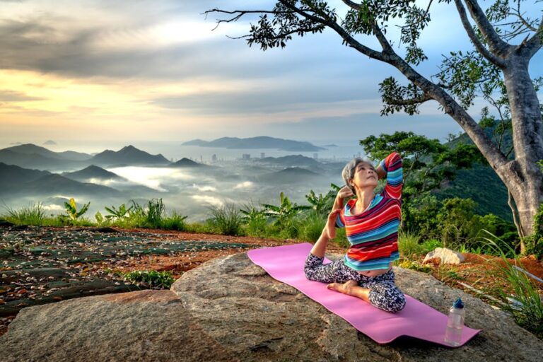 Senior woman practices yoga outdoors on a mountain at sunset, embracing wellness and serenity.