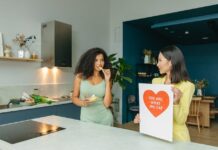 7 Hábitos saudáveis que transformam sua vida Two women discussing nutrition in a kitchen, holding health-related posters.