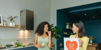 7 Hábitos saudáveis que transformam sua vida Two women discussing nutrition in a kitchen, holding health-related posters.