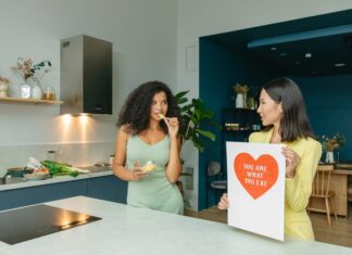 7 Hábitos saudáveis que transformam sua vida Two women discussing nutrition in a kitchen, holding health-related posters.