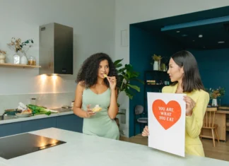 7 Hábitos saudáveis que transformam sua vida Two women discussing nutrition in a kitchen, holding health-related posters.