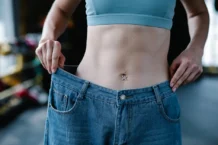 Exercícios para perder barriga em casa: Resultados em 21 dias A woman shows her weight loss by holding oversized jeans revealing her toned stomach.