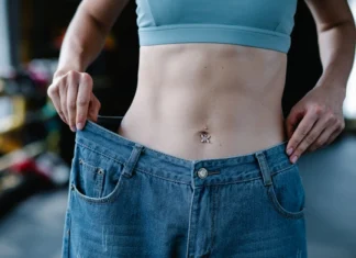 Exercícios para perder barriga em casa: Resultados em 21 dias A woman shows her weight loss by holding oversized jeans revealing her toned stomach.