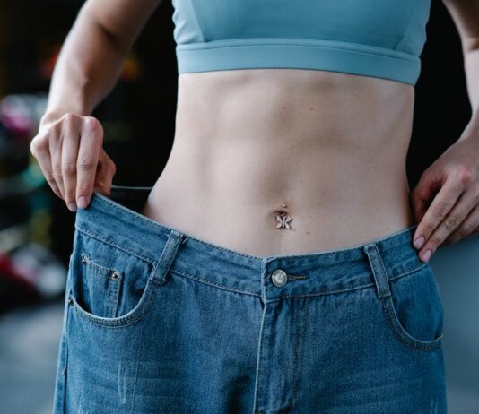 Exercícios para perder barriga em casa: Resultados em 21 dias A woman shows her weight loss by holding oversized jeans revealing her toned stomach.