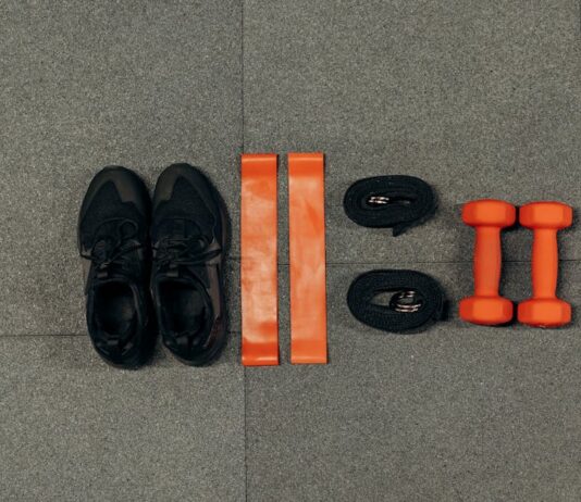 Treino simples para perder peso Flat lay of gym essentials including dumbbells, sneakers, and resistance bands on a grey floor.