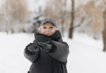 Elderly woman wearing a winter jacket and cap stretches arms outdoors in a snowy park.