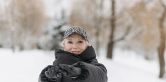 Elderly woman wearing a winter jacket and cap stretches arms outdoors in a snowy park.