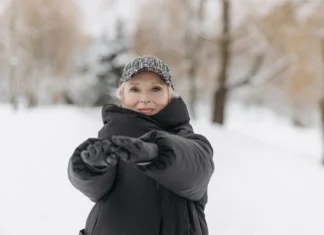 Como manter a forma no inverno: Dicas surpreendentes Elderly woman wearing a winter jacket and cap stretches arms outdoors in a snowy park.