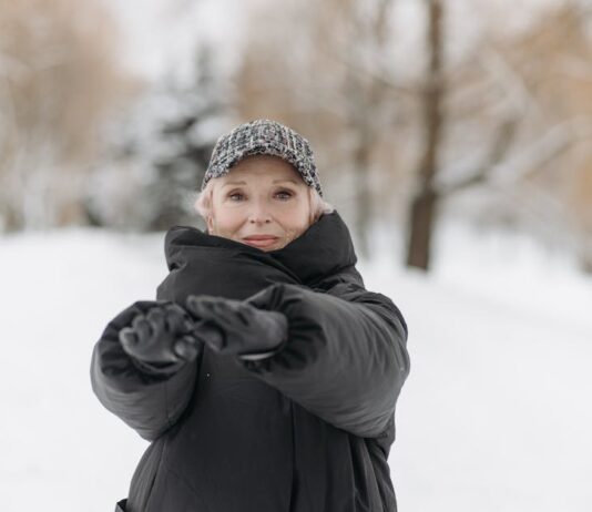Como manter a forma no inverno: Dicas surpreendentes Elderly woman wearing a winter jacket and cap stretches arms outdoors in a snowy park.