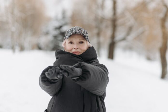 Como Manter a Forma no Inverno: Dicas Surpreendentes! Elderly woman wearing a winter jacket and cap stretches arms outdoors in a snowy park.