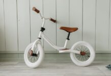 Descubra as melhores ciclovias para iniciantes no Brasil A white kids' balance bike with brown accents standing indoors against a wooden wall.
