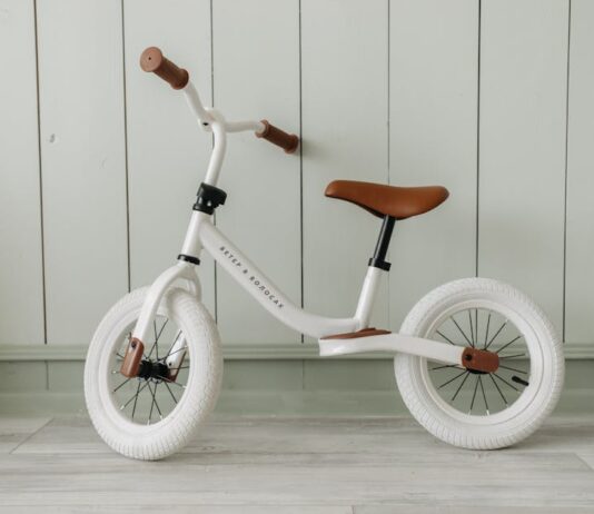 Descubra as melhores ciclovias para iniciantes no Brasil A white kids' balance bike with brown accents standing indoors against a wooden wall.
