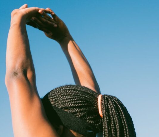Dieta e exercício antes do verão: Surpreenda-se com os resultados Person stretching outdoors with braided hair against a clear blue sky for a healthy lifestyle concept.